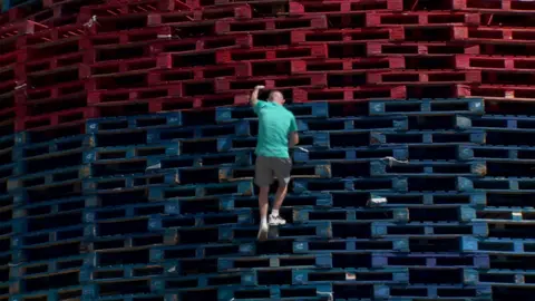 a man climbing a stack of red and blue pallets - he is wearing a T shirt, shorts and trainers