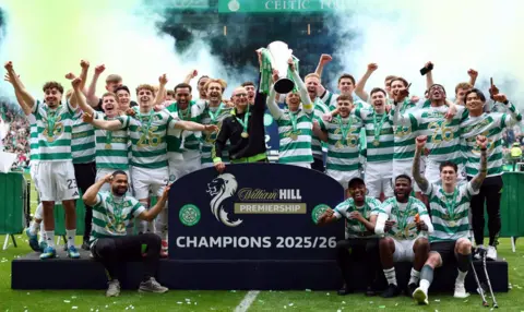 Reuters Celtic players celebrate winning the Scottish Premiership title at Celtic Park, lifting the trophy and posing for a team photo in front of a “Champions 2025/26” sign.