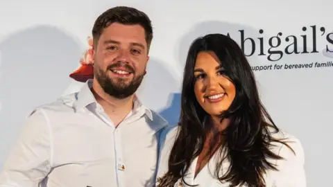 Matt Knight on the left has dark short hair and a short beard, he is wearing a white shirt. Hannah Spearing next to his on the right has long dark hair and is wearing a white blazer. They're both smiling.