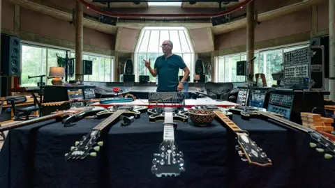 Yorktillyer A man is standing behind several electric guitars laid out on a table, in a large studio building with many electronics desks behind him.
