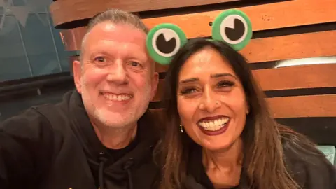LNR Steve and Payal, smiling and looking at the camera, with wooden slates behind them. Steve has short grey hair and a beard, wearing a black top, Payal, has on green frog ears, has long dark hair, a nose piercing, earrings and a black top. 
