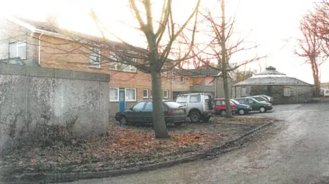 West Yorkshire Police A red-brick building with a blue door and several large windows. Next to it, several cars are parked and there are skeletal trees standing next to the cars, with leaves on the ground underneath them. In the rear of the picture is a round building with a low pointed roof.