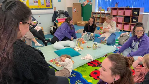 A group of women sit in a circle in a room filled with children's toys. Some are holding their children. Some are wearing purple tops.