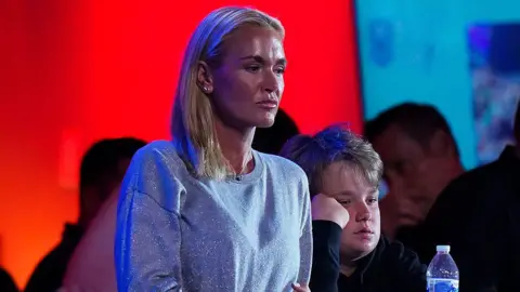A woman with blonde hair looks ahead seriously. Her hair is all pulled to the right side of her shoulder. She is wearing a silver sparkly sweater and a pendant necklace. There are people in the background.