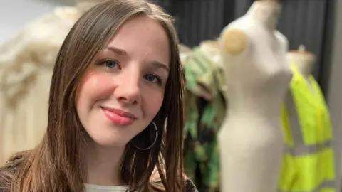 Scarlett Baker has long brown hair and is wearing a textured brown cardigan over a light top. Behind her, there are some mannequins, one features a cream‑coloured dress, another has a green patterned outfit, and a third is dressed in a bright yellow high‑visibility jacket but they are not in focus. 