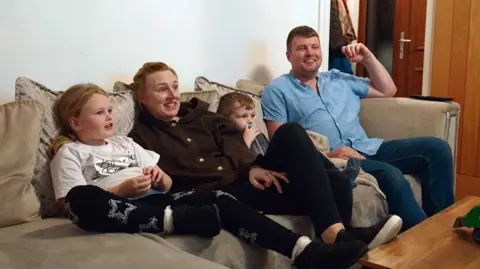 Two children sit on a grey sofa smiling alongside their mum and dad.