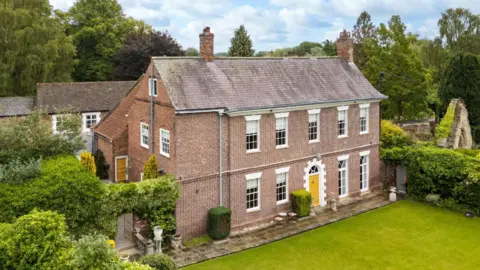 A large stone detached house with surrounded by green hedges and lawns