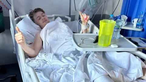 Family A young man laying in a hospital bed. He has white sheet over him and has two thumbs up in the air. He has tubes attached to his left hand.