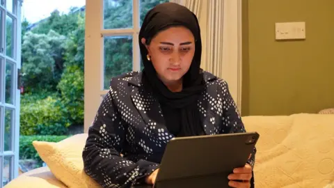 Salma Niazi Salma Niazi looks at an electronic device. She is wearing a blazer and a black head covering. She is sat on a couch in front of an open door. Outside, there are trees.