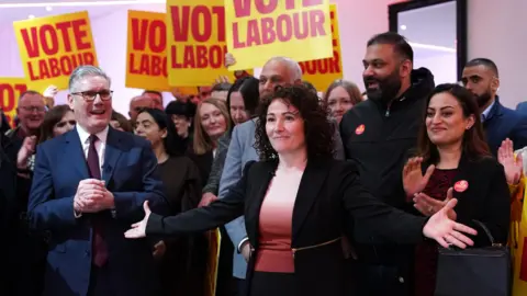 Getty Images Labour by-election candidate Angeliki Stogia speaks alongside Prime Minister Keir Starmer as they campaign ahead of the Gorton and Denton by-election at Rushford Park sports complex. She holds out her arms widely as Sir Keir claps at the side.