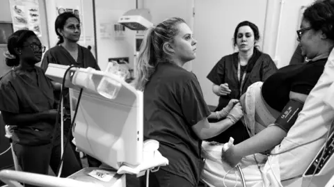 James Clifford Kent A group of four midwives in dark scrubs gathered around a medical monitoring station, looking focused as they review data on a screen in a bright clinical room.