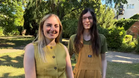 Amy Holmes/BBC Two women standing in a garden with mature trees, lawns and buildings visible in the background in St Albans. Both are smiling.