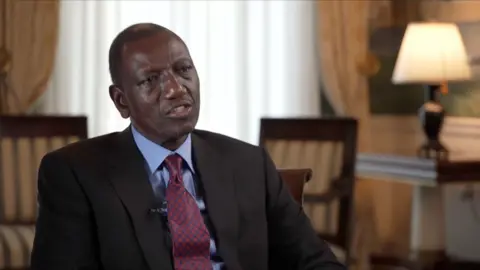 President of Kenya William Ruto wearing a suit with a plaid burgundy tie seated on an elegant room