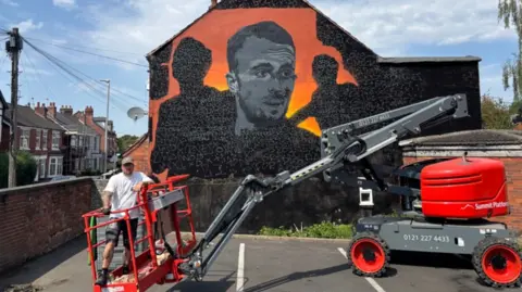 The mural shows a painting of a man looking to the right in black and white on an orange background. The artist, a man, is wearing a baseball cap and standing in the red canopy of a machine to raise him up in the air.