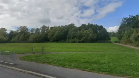 Google A large open playing field with a concrete path running through it and a large copse of trees 