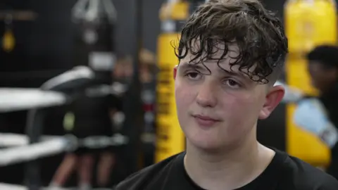 George looks away from the camera. He is sweating from exercise and has short curly brown hair and is wearing a black t-shirt.