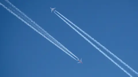 Two planes high in the blue sky pass each other. Both have two white contrails in their wake.