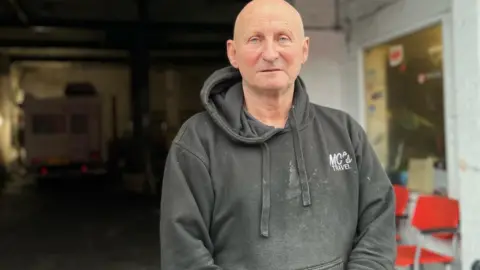 Jonny Humphries/BBC Paul Jones in his 60s, who has a bald head and blue eyes and is wearing a black hooded sweatshirt, looks into the camera with his garage workshop in the backdrop.