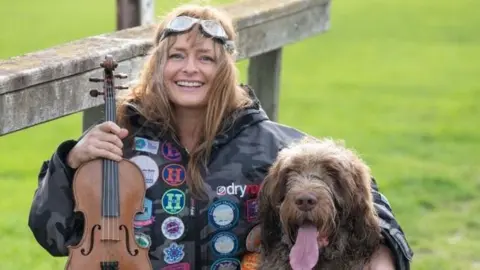 Laura Reineke Laura is holding a violin whilst her other hand is on her brown dog with it's tongue sticked out. 