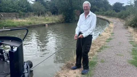 Kate Bradbrook/BBC A man standing at a canal towpath wearing a white shirt, black trousers, and holding a rope, connected to a boat. He has short grey hair and is smiling. Trees and foliage is all around the canal. The path has weeds on it. 