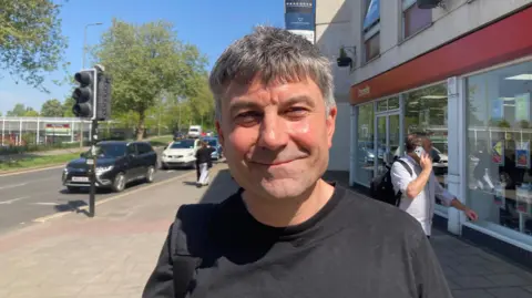 Maurice East smiles for the camera outside a shopping centre. He is wearing a black t-shirt and has short, straight black hair with grey at the sides. Cars and a pedestrian can be seen in the background.