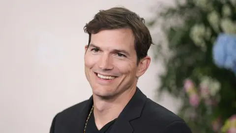Getty Images A head and shoulders shot of Ashton Kutcher in which he looks at the camera and smiles. he has on a dark jacket and a gold chain around his neck. 