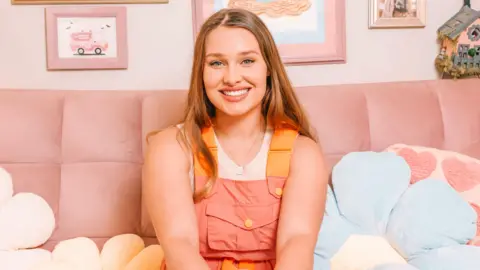 Tesni DeLuna, who has long wavy light brown hair and is wearing a white t-shirt with pink and orange dungarees over the top and smiling looking at the camera. She is sat on a pink sofa bed, with blue, pink and yellow flower and heart cushions on either side of her, with a pink background and various paintings and frames pinned up on the wall.