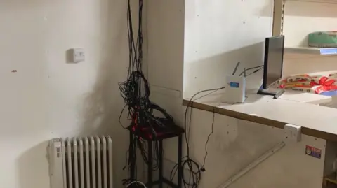 The corner of a room with lots of wires in a bundle and hanging down from the ceiling, next to a small radiator, where CCTV had been installed but has since been stolen.