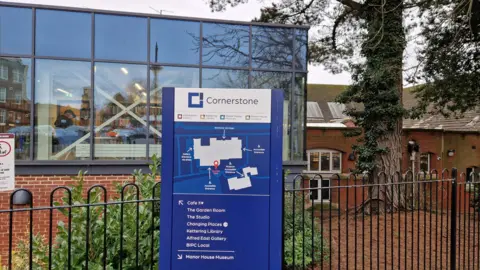 Pete Cooper/BBC A single-storey red brick building with a large window revealing storage shelves within. A blue sign outside says "Cornerstone" and shows a map of the site. There are railings behind the sign.