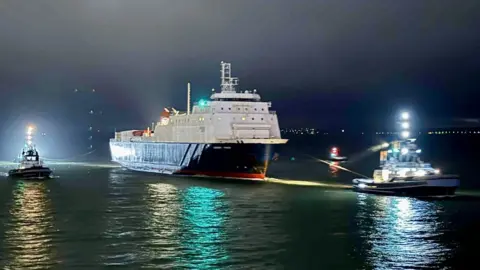 The dark blue and white Caesarea Trader in Portsmouth Harbour being towed by two tugs