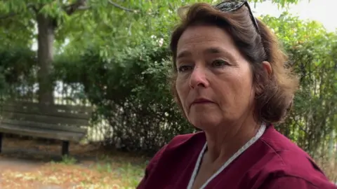 A woman wearing purpley red scrubs sits in a park. She is probably in her late 50s or early 60s and has short brown hair, She has sunglasses on top of her head.