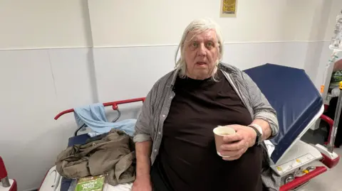 Patient waiting on a hospital trolley in A&E with a paper cup of tea in his hand.