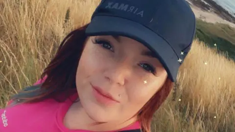 Facebook Woman - Lucretia Donaghy - smiling at camera, wearing a pink top and a dark blue baseball cap.