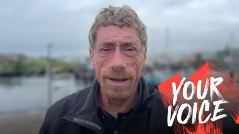 BBC Male with gray hair and ginger beard with a navy jacket looking at the camera. Blurred background at a pier.