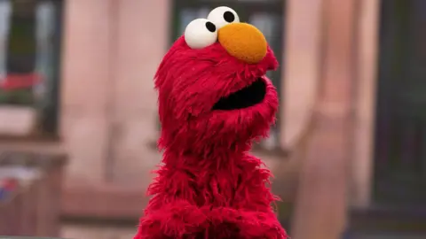 Elmo on the Sesame Street set. Elmo is a furry red Muppet with a large orange nose and big round eyes. He looks up to the right of the frame, with the set blurred behind him. 