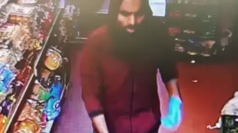 West Yorkshire Police A blurry image of a man with a red hooded top on and the hood up over his head. He has dark eyes and a dark beard.