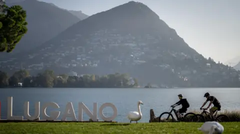 FABRICE COFFRINI/AFP via Getty Images Swans are seen next to a sign on the shores of Lake Lugano in Lugano, southern Switzerland. Two people are cycling near the lake.