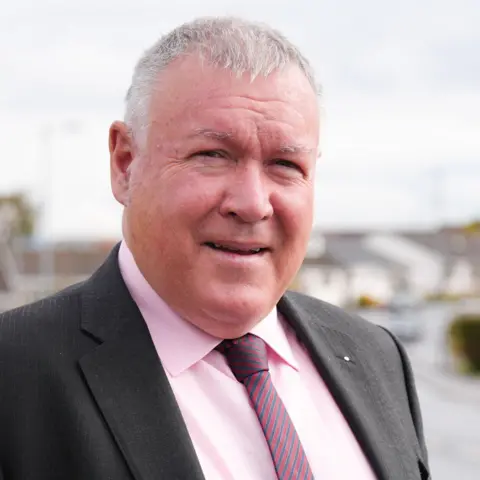 Scottish Labour Party A man with short grey hair, wearing a dark coloured suit, a pink shirt and a striped tie. Houses can be seen in the background.