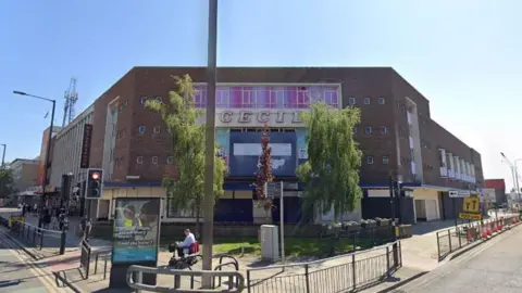 Google A large brown building located at the corner of a road in a city, near traffic lights. It has several small square windows and says 'CECIL' across the middle of it in bold writing beneath a long window.