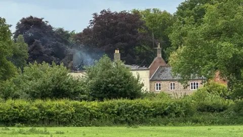Andrew Turner/BBC Building behind some trees and shrubbery with gutted roof. Smoke can be seen from the roof.