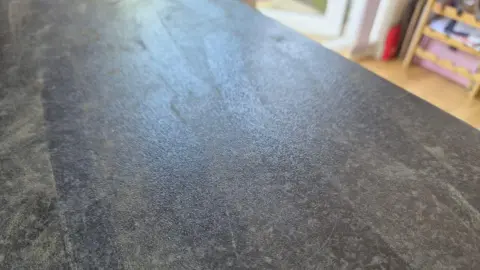 Kayleigh Paterson A black kitchen counter which has some dust on it. You can see the floor in the top right of the image with some wooden shelves.