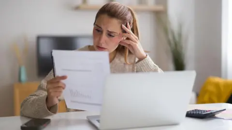 A woman sat at a table looking at a piece of paper that looks like a bill, sat in front of a calculator and a laptop computer, looking concerned.