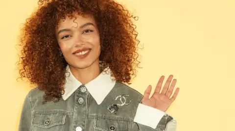 Anthony Kwan / Getty Images Whitney Peak, with long curly hair and wearing a grey jean jacket with a Chanel logo broach pinned to it, waves - Tuesday 5 November 2024