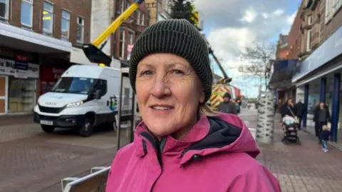 A woman in a dark woollen hat, wearing a pink coat. She is smiling as she stands in front of the Christmas tree. A council cherry-picker van is parked alongside the tree.