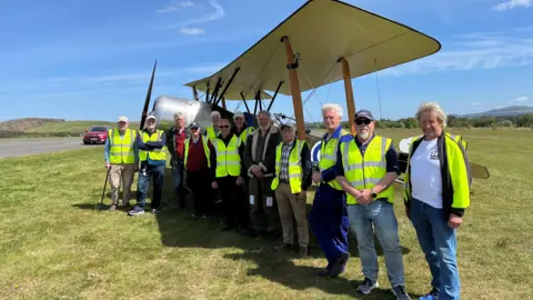 The Sopwith Strutter, a World War One small biplane, sits proudly on the grass at RAF Kirknewton, with all of those who worked on the project lined up along the wingspan, mainly mature gentlemen in yello hi-vis vests.
