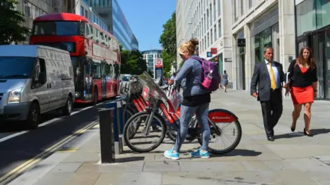 A woman is hiring a Santander Cycles bicycle on the pavement, with her hands on both handlebars. She is standing up to the left of the bike, and has blonde hair and is dressed in striped grey-blue leggings and long-sleeve top, with a purple backpack. The bicycle is parked in a docking station, and is at the front of a row of bikes parked on the pavement alongside a road. Traffic is on the left, flowing next to the bikes. A man and a woman are walking past on the right, and the street and buildings are visible in the background.
