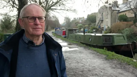 A man is stood on the side of a canal, he is wearing black framed glasses and a navy jacket and jumper. Behind him you can see a number of narrow boats. 