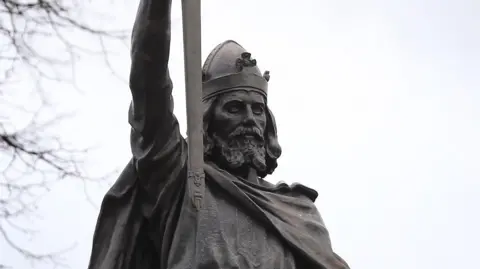 A statue of King Alfred the Great. He is wearing a helmet and holding a sword in the air. 