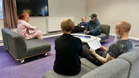 Martin Giles/BBC Five people sitting on sofas facing one another. One is wearing a pink tracksuit and has red hair, another is wearing a black dress with short blonde hair. A bald man with a beard sits reading a piece of paper. Another man is reading a book, another wears a jumper and baseball cap. 