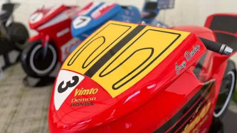 A close up of a red sculpture which bears the number 3, and name Joey Dunlop. You can see three other colourful models in the background